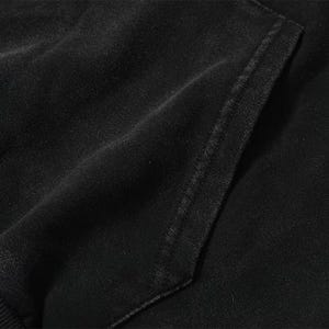 May include: Close-up of a black hoodie pocket. The fabric appears to be a soft, textured material. The pocket is rectangular and stitched with a contrasting thread. The overall aesthetic is casual and comfortable.