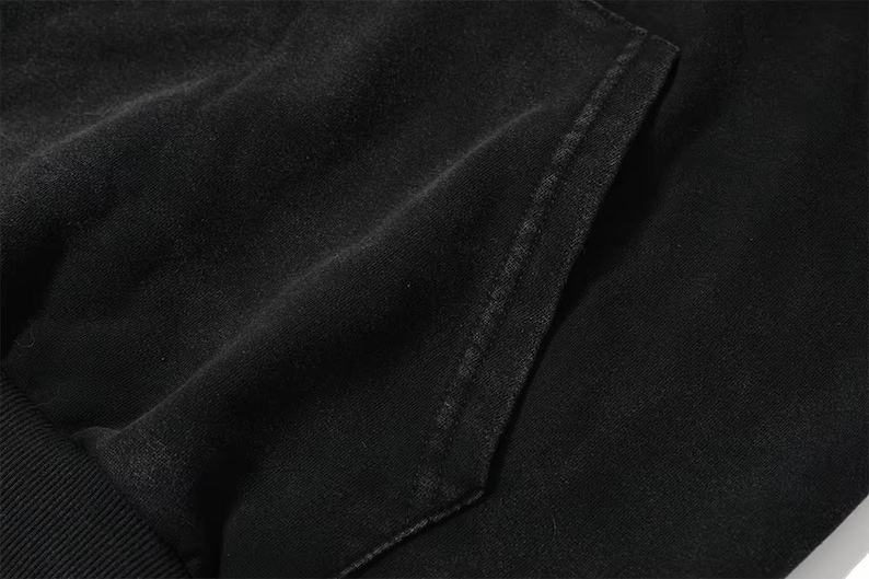 May include: Close-up of a black hoodie pocket. The fabric appears to be a soft, textured material. The pocket is rectangular and stitched with a contrasting thread. The overall aesthetic is casual and comfortable.