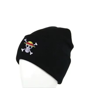 May include: Black beanie hat with a white skull and crossbones logo featuring a yellow straw hat. The logo is embroidered on the front of the hat. The hat is made of a soft, knitted material.