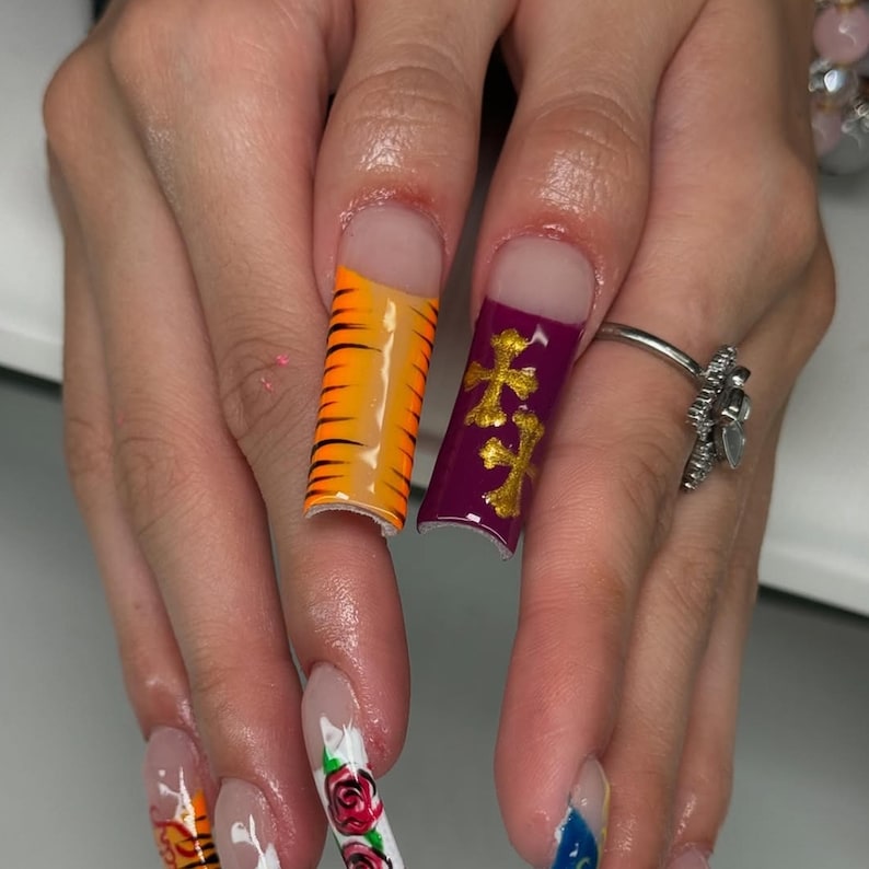 May include: Close-up of long, decorated fingernails. One nail features orange and black tiger stripes, while another displays gold crosses on a purple background. Other nails have floral and abstract designs. A silver ring with a floral design is also visible.
