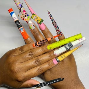 Puede incluir: Uñas largas y decoradas con temática escolar. Los diseños incluyen una barra de pegamento, una paleta de pintura, un crayón, una regla, un cuaderno, un lápiz y una uña cubierta de pedrería de colores. Las uñas están pintadas en varios colores, incluyendo rosa, blanco y amarillo.