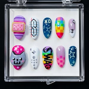 May include: A set of ten artificial nails with various designs. The nails feature colorful patterns, including stripes, a pixelated cat, and the word "SWAG." Other designs include stars, a rainbow, and the text "XD" and ":3."