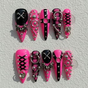 May include: A set of press-on nails in a variety of designs. The nails are primarily hot pink and black, with silver-colored metal accents. Some nails feature an X design, while others have metal rings and studs. The nails are pointed.