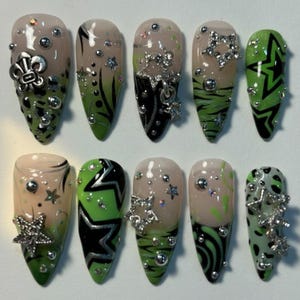 May include: A set of ten artificial nails with various designs. The nails feature a combination of green, black, and nude colors, adorned with silver embellishments like stars, beads, and a fleur-de-lis. Some nails have geometric patterns and animal print.