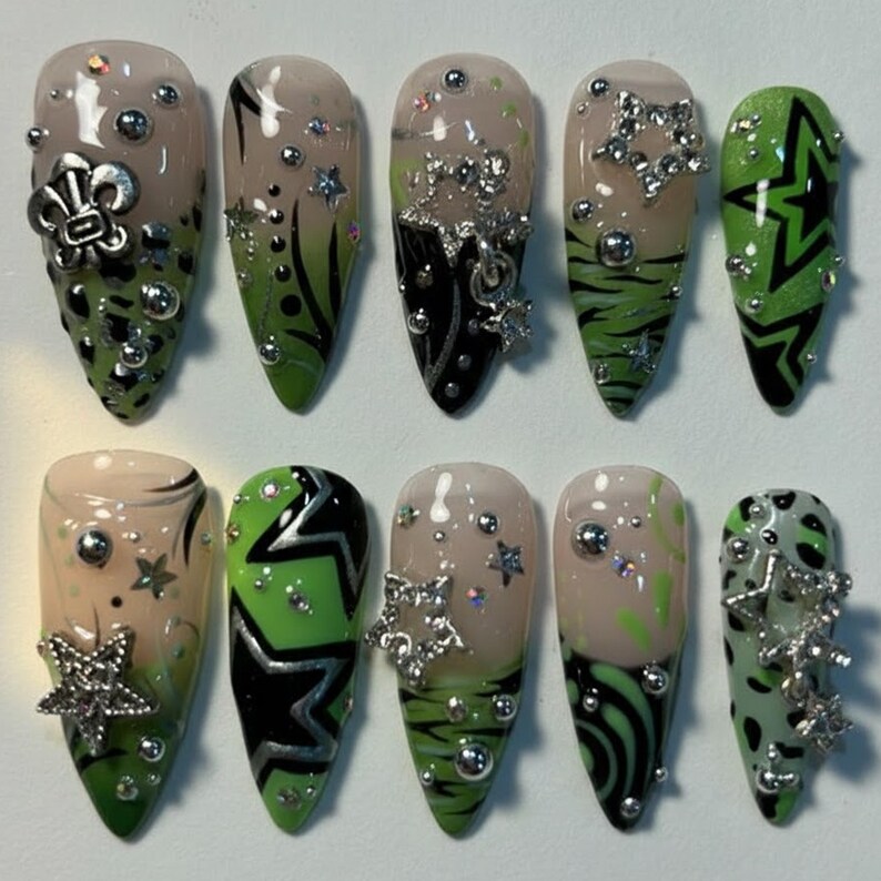 May include: A set of ten artificial nails with various designs. The nails feature a combination of green, black, and nude colors, adorned with silver embellishments like stars, beads, and a fleur-de-lis. Some nails have geometric patterns and animal print.