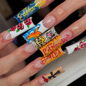 May include: Long, square acrylic nails with diverse designs. The nails have a French tip design with artwork including roses, a koi carp, a tiger, and the text "Ed Hardy" in red and gold. One nail features a cross and a red bead.