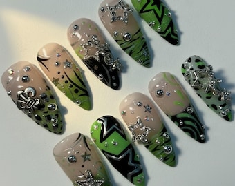 May include: A set of ten long, almond-shaped artificial nails. The nails feature a variety of designs, including green and black star patterns, silver star and stud embellishments, and green and black tiger stripes. Some nails have a nude base.