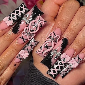 May include: A set of long, artificial nails with a pink and black design. The nails feature pink bows, silver embellishments, and a criss-cross pattern. The design is intricate and detailed, with a glossy finish.