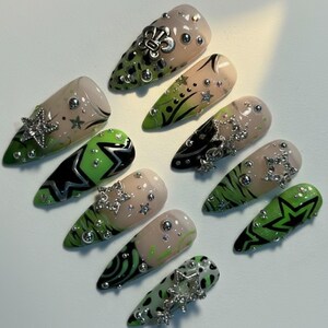 May include: A collection of almond-shaped, artificial nails. The nails feature a variety of designs, including green and black patterns, silver star and floral embellishments, and clear rhinestones. Some nails have a nude base with green tips.