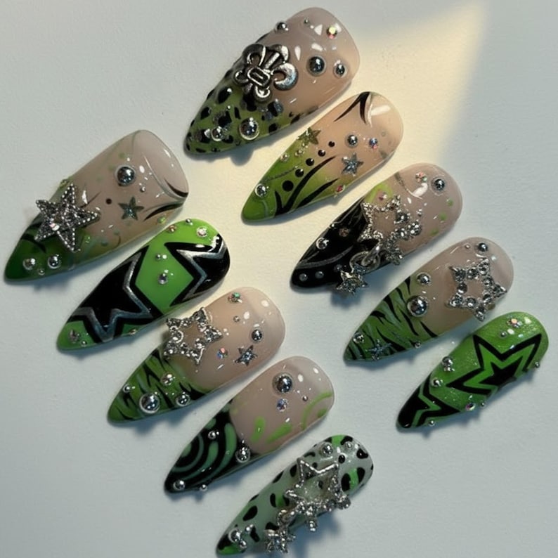 May include: A collection of almond-shaped, artificial nails. The nails feature a variety of designs, including green and black patterns, silver star and floral embellishments, and clear rhinestones. Some nails have a nude base with green tips.