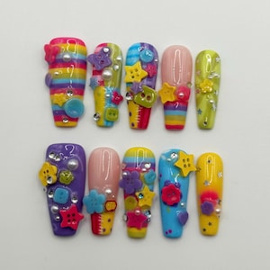 May include: A collection of ten artificial nails, each uniquely decorated. Designs include stripes, buttons, stars, and pearls. The nails are painted in a variety of colours, including pink, yellow, blue, and purple, for a vibrant look.