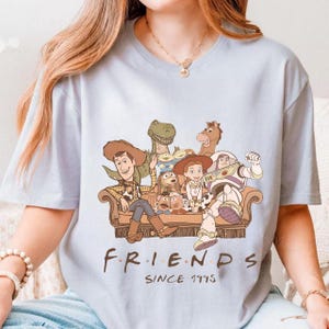 May include: Light blue t-shirt featuring a cartoon illustration of Toy Story characters seated on a brown couch. The word "FRIENDS" is printed below the image, with "SINCE 1995" underneath.