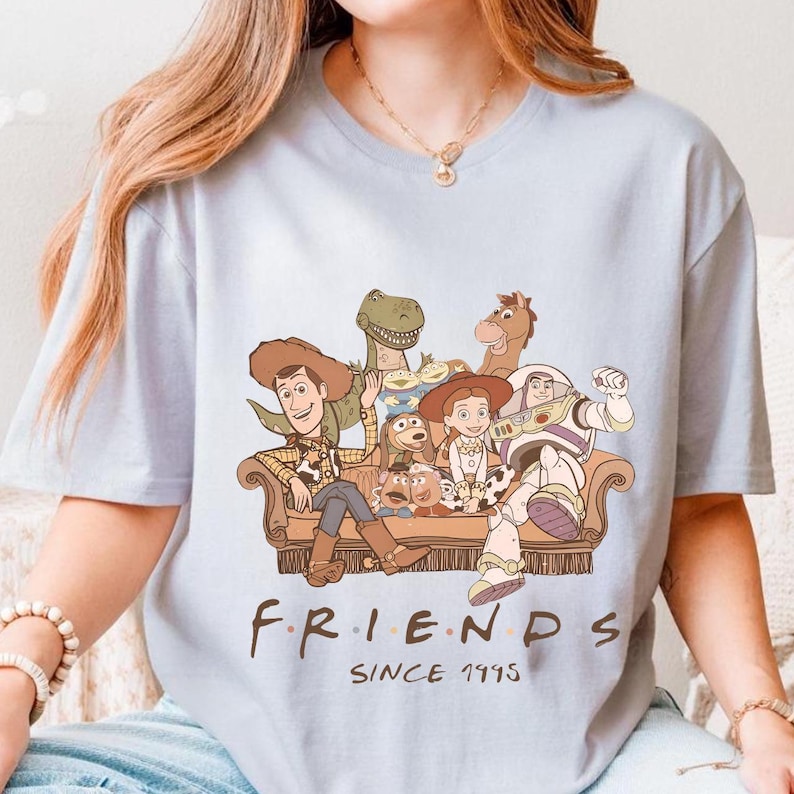 May include: Light blue t-shirt featuring a cartoon illustration of Toy Story characters seated on a brown couch. The word "FRIENDS" is printed below the image, with "SINCE 1995" underneath.