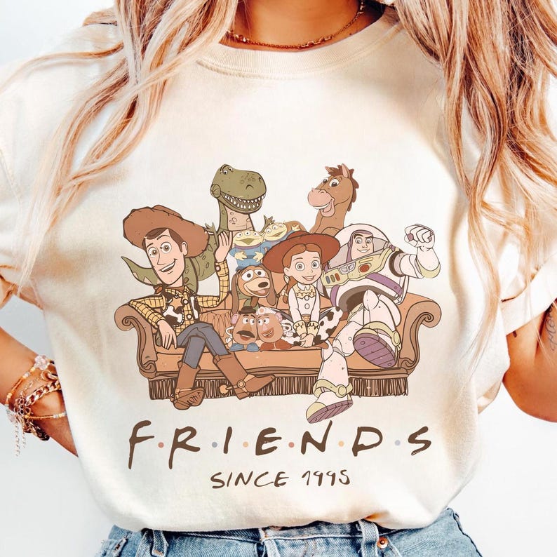May include: Cream-colored t-shirt featuring a graphic of Toy Story characters sitting on a couch. The characters include Woody, Buzz Lightyear, Rex, Jessie, and others. The word "FRIENDS" is printed below the image, with "SINCE 1995" underneath.