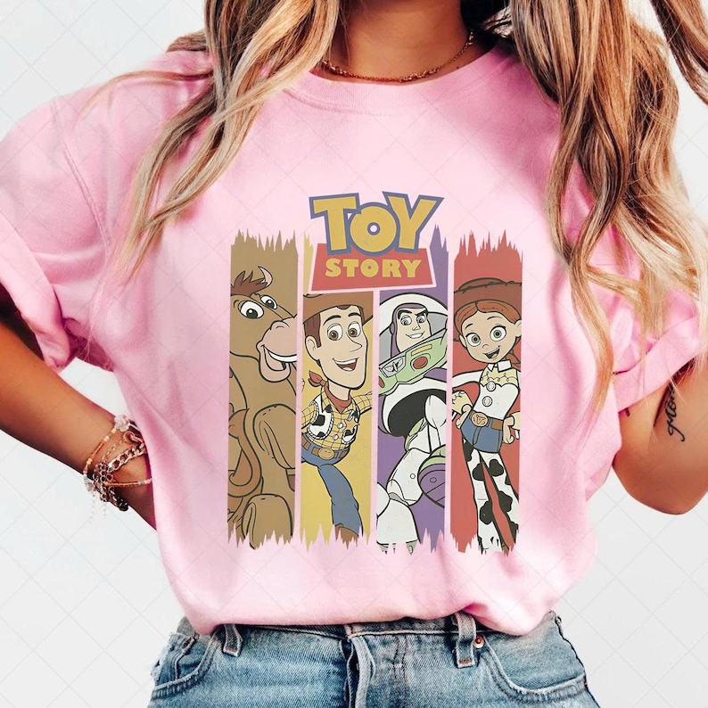 May include: A pale pink t-shirt with the words "TOY STORY" in yellow and characters from the film. The characters, including a brown horse, Woody, Buzz Lightyear, and Jessie, are displayed in vertical stripes.