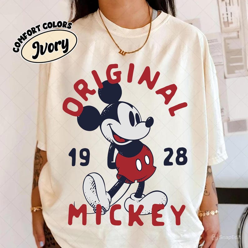 May include: Ivory t-shirt with a Mickey Mouse graphic. The design features "ORIGINAL MICKEY 1928" in red, alongside Mickey in red shorts and white gloves. The shirt is labelled "COMFORT COLORS". The t-shirt is a cream colour.