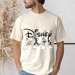 May include: Cream-colored t-shirt featuring the Disney logo and a graphic of Disney characters crossing a street. The graphic includes Goofy, Donald Duck, and Mickey Mouse, with Pluto. The word "Disney" is in black script.