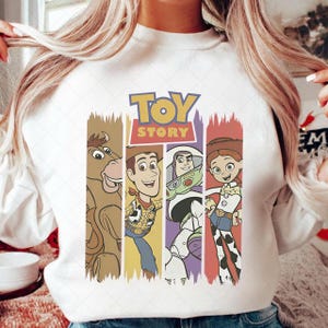 May include: White sweatshirt with a vibrant graphic of characters from the animated film "Toy Story." The design includes Woody, Buzz Lightyear, Jessie, and Bullseye, with the "Toy Story" logo at the top.
