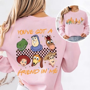 May include: Pink sweatshirt featuring cartoon character faces and the text "YOU'VE GOT A FRIEND IN ME." The design includes a checkered pattern and star accents. The sweatshirt has long sleeves and a crew neck.