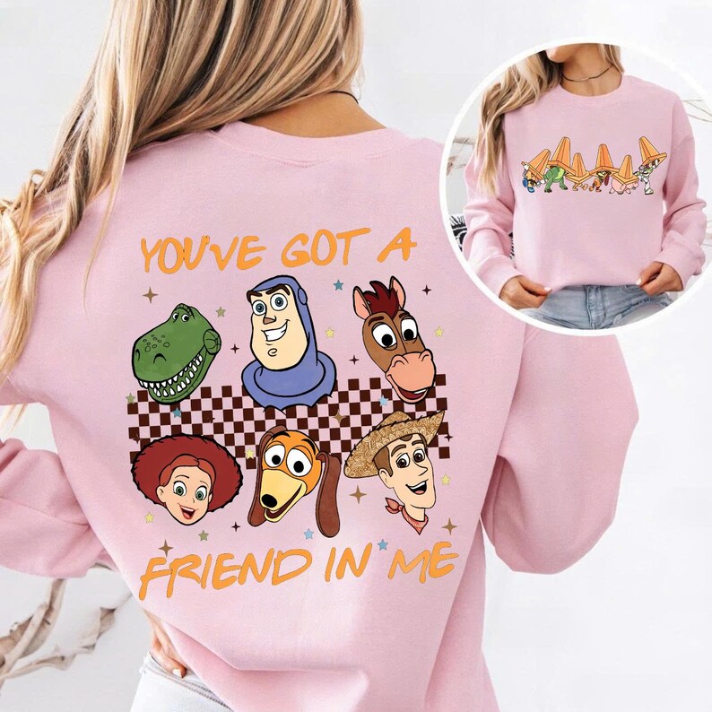 May include: Pink sweatshirt featuring cartoon character faces and the text "YOU'VE GOT A FRIEND IN ME." The design includes a checkered pattern and star accents. The sweatshirt has long sleeves and a crew neck.