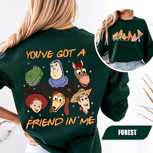 May include: Forest green sweatshirt featuring cartoon character graphics and the text "YOU'VE GOT A FRIEND IN ME". The design includes images of a dinosaur, Buzz Lightyear, a horse, Jessie, Slinky Dog, and Woody.