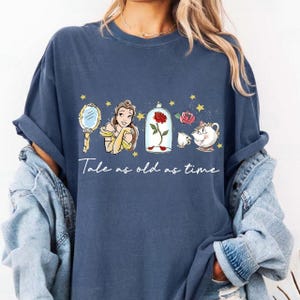 Puede incluir: Una camiseta azul marino con un diseño de La Bella y la Bestia. El diseño incluye a Bella, un espejo, una rosa en una campana de cristal y el texto "Tale as old as time."