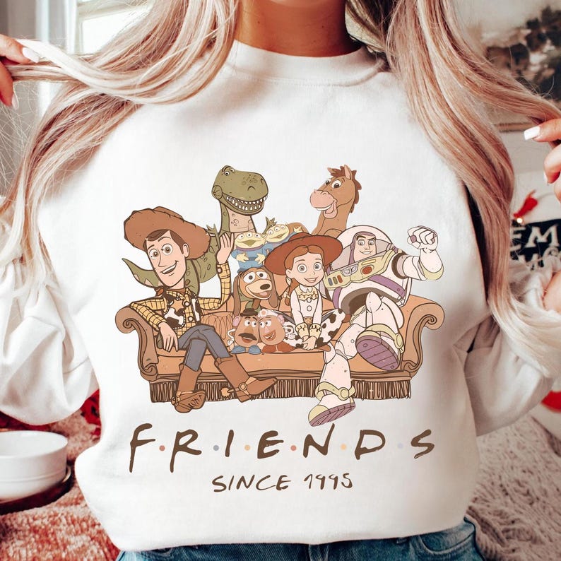 May include: White sweatshirt featuring a graphic of Toy Story characters on a brown couch. The word "FRIENDS" is written in a stylized font, with "SINCE 1995" below. The design includes Woody, Buzz Lightyear, and other characters.