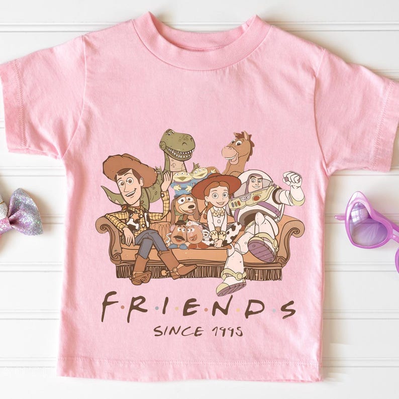 May include: A pink t-shirt featuring a graphic of characters from the movie Toy Story, including Woody, Buzz Lightyear, and Jessie, seated on a brown couch. The word "FRIENDS" is printed below the graphic, with "SINCE 1995" underneath.