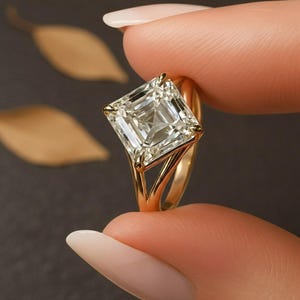 May include: A gold ring featuring a large, square-cut diamond. The diamond is clear and reflects light, set in a gold band with a split shank design. The ring is held up against a dark background.