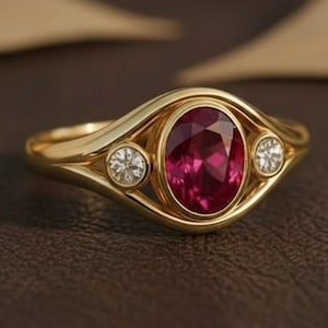 May include: A gold ring featuring an oval, deep red gemstone as the centerpiece, flanked by two smaller, round, clear gemstones. The ring's band has a unique, flowing design. The ring is set against a brown background.