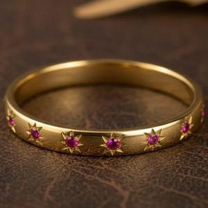 May include: A gold-colored ring with a series of small, star-shaped designs, each containing a small, red gemstone. The ring is set against a brown leather-like surface, with blurred leaves in the background. Jewelry.