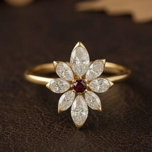 Könnte beinhalten: Ein goldener Ring mit einem floralen Design. Die Blume besteht aus weißen, marquiseförmigen Edelsteinen, die einen zentralen roten Edelstein umgeben. Der Ring ist vor einem dunkelbraunen Hintergrund mit verschwommenen Blättern im Hintergrund platziert.