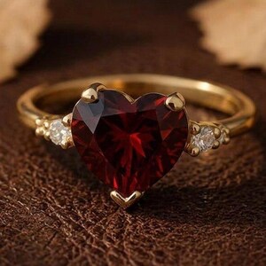 May include: A gold ring featuring a large, heart-shaped, deep red gemstone as the centerpiece. The band is accented on each side by a small, clear, round-cut stone. The ring is crafted with a polished gold finish and a classic prong setting.