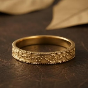 Puede incluir: Un anillo de oro con un diseño floral. El anillo presenta un patrón repetido de flores y vides, bordeado por una línea punteada. Se encuentra sobre un fondo marrón oscuro, con dos hojas borrosas.