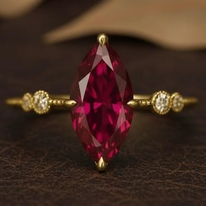 以下が含まれることがあります： 大きなマーキス型のルビーの宝石をあしらったゴールドリング。ルビーは深い赤色で、両側に小さな丸いダイヤモンドが付いています。リングは濃い茶色の背景にセットされています。
