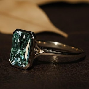 Puede incluir: Un anillo de plata con una gran piedra preciosa rectangular de color verde azulado. La piedra preciosa está engastada en un bisel. El anillo está sobre una superficie marrón oscuro, con hojas marrones borrosas en el fondo. Es una joya.