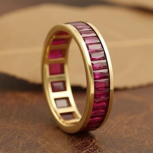 Könnte beinhalten: Ein goldener Ring mit einem Band aus rechteckigen, tiefroten Edelsteinen. Der Ring ist vor einem braunen Hintergrund platziert, der den Kontrast zwischen dem Gold und den roten Steinen hervorhebt. Der Ring ist ein Schmuckstück.