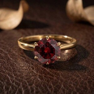 Könnte beinhalten: Ein Goldring mit einem runden, tiefroten Edelstein in einer Fassung mit sechs Krappen. Die Ringschiene ist glatt und poliert und zeigt ein klassisches Design, das die leuchtende Farbe und den Facettenschliff des zentralen Steins hervorhebt.