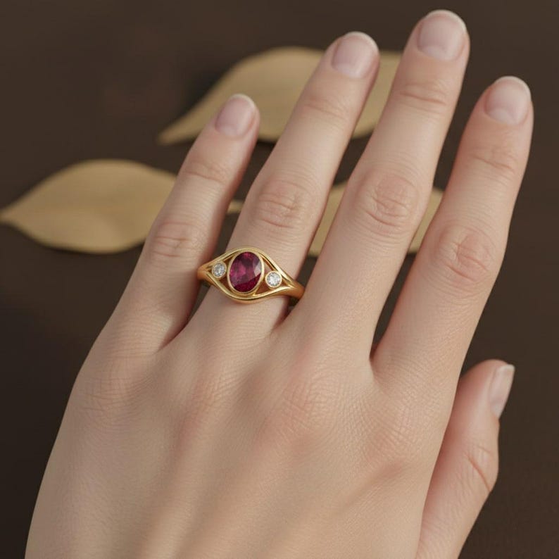 Puede incluir: Un anillo de oro con un gran rub&iacute; ovalado en el centro, flanqueado por dos piedras preciosas claras y redondas m&aacute;s peque&ntilde;as. El anillo tiene una banda de oro con un dise&ntilde;o calado delicado. El anillo est&aacute; en un dedo.
