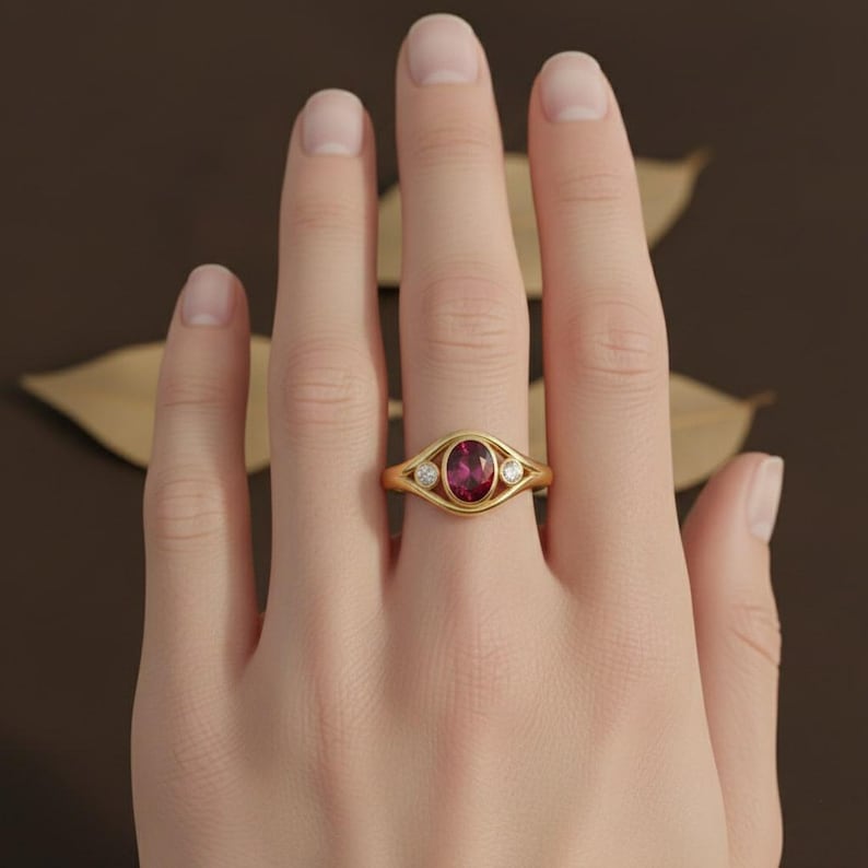 Puede incluir: Un anillo de oro con una piedra central ovalada de color rojo intenso, flanqueada por dos piedras m&aacute;s peque&ntilde;as, redondas y transparentes. La banda del anillo tiene un dise&ntilde;o delicado y abierto. El anillo se muestra en una mano.