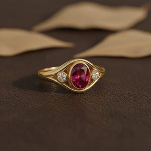 Puede incluir: Un anillo de oro con una piedra preciosa roja oscura ovalada en el centro, flanqueada por dos piedras preciosas redondas y claras m&aacute;s peque&ntilde;as. El anillo tiene un dise&ntilde;o &uacute;nico y fluido. El anillo est&aacute; sobre un fondo marr&oacute;n.