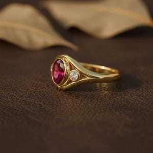 Puede incluir: Un anillo de oro con una gran gema de rub&iacute; ovalada y dos diamantes redondos m&aacute;s peque&ntilde;os. El anillo tiene una banda lisa y pulida y un dise&ntilde;o cl&aacute;sico y elegante. Las piedras preciosas est&aacute;n engastadas en un engaste de oro.
