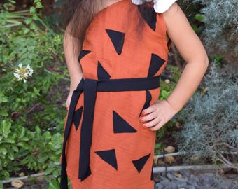 Könnte beinhalten: Ein Kind in einem handgefertigten orangefarbenen und schwarzen Kostüm, das an die Kleidung eines Höhlenbewohners erinnert. Das Kleid hat einen gezackten Saum und einen schwarzen Gürtel. Das Kind hat lange, dunkle Haare und steht draußen.