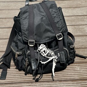 May include: Black nylon backpack with multiple zippered pockets, adjustable straps, and silver-tone hardware. A white and black dinosaur skeleton charm hangs from the front. The backpack has a flap closure.