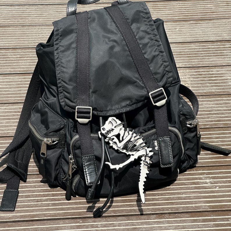 May include: Black nylon backpack with multiple zippered pockets, adjustable straps, and silver-tone hardware. A white and black dinosaur skeleton charm hangs from the front. The backpack has a flap closure.