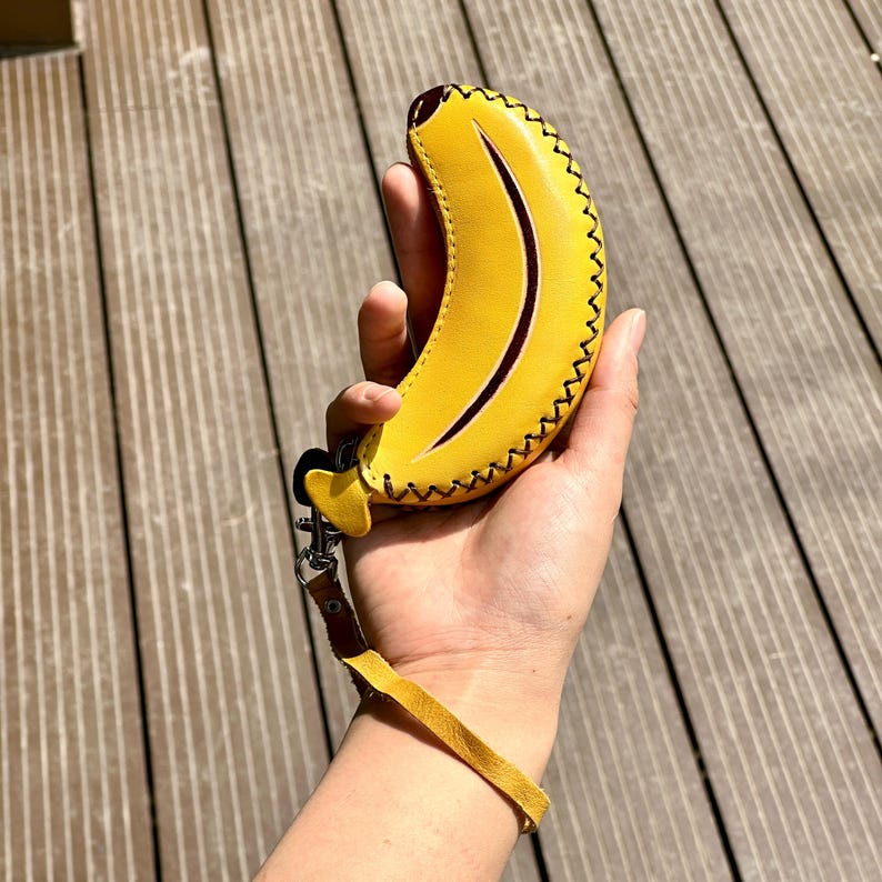 K&ouml;nnte beinhalten: Ein gelber, lederner, bananenf&ouml;rmiger Schl&uuml;sselanh&auml;nger mit schwarzen N&auml;hten und einem braunen Akzent. Der Schl&uuml;sselanh&auml;nger hat eine braune Handschlaufe und einen silbernen Verschluss. Das Bananen-Design ist mit einer dunkelbraunen, gebogenen Linie versehen.
