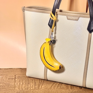K&ouml;nnte beinhalten: Eine cremefarbene Tragetasche mit schwarzen und beigen Griffen, mit einem gelben, bananenf&ouml;rmigen Schl&uuml;sselanh&auml;nger. Der Schl&uuml;sselanh&auml;nger hat einen Rei&szlig;verschluss und eine schwarze Kontur. Die Tasche hat eine strukturierte Oberfl&auml;che und eine hellbeige Umrandung.