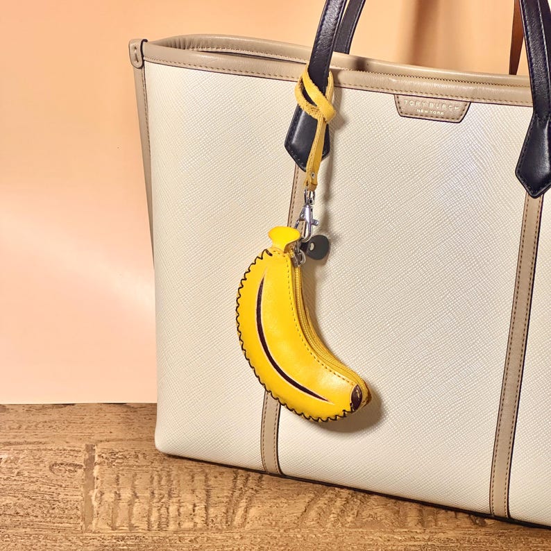 K&ouml;nnte beinhalten: Eine cremefarbene Tragetasche mit schwarzen und beigen Griffen, mit einem gelben, bananenf&ouml;rmigen Schl&uuml;sselanh&auml;nger. Der Schl&uuml;sselanh&auml;nger hat einen Rei&szlig;verschluss und eine schwarze Kontur. Die Tasche hat eine strukturierte Oberfl&auml;che und eine hellbeige Umrandung.