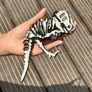 May include: A white and black dinosaur skeleton made of leather is held in a hand. The dinosaur has a detailed design with black outlines and accents. The skeleton is a unique decorative item.