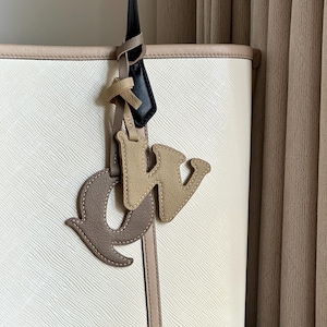May include: Cream tote bag with a textured surface and beige trim. A leather charm hangs from the strap, featuring the letters "CW" in brown and beige, stitched with contrasting thread. The bag's strap is black.